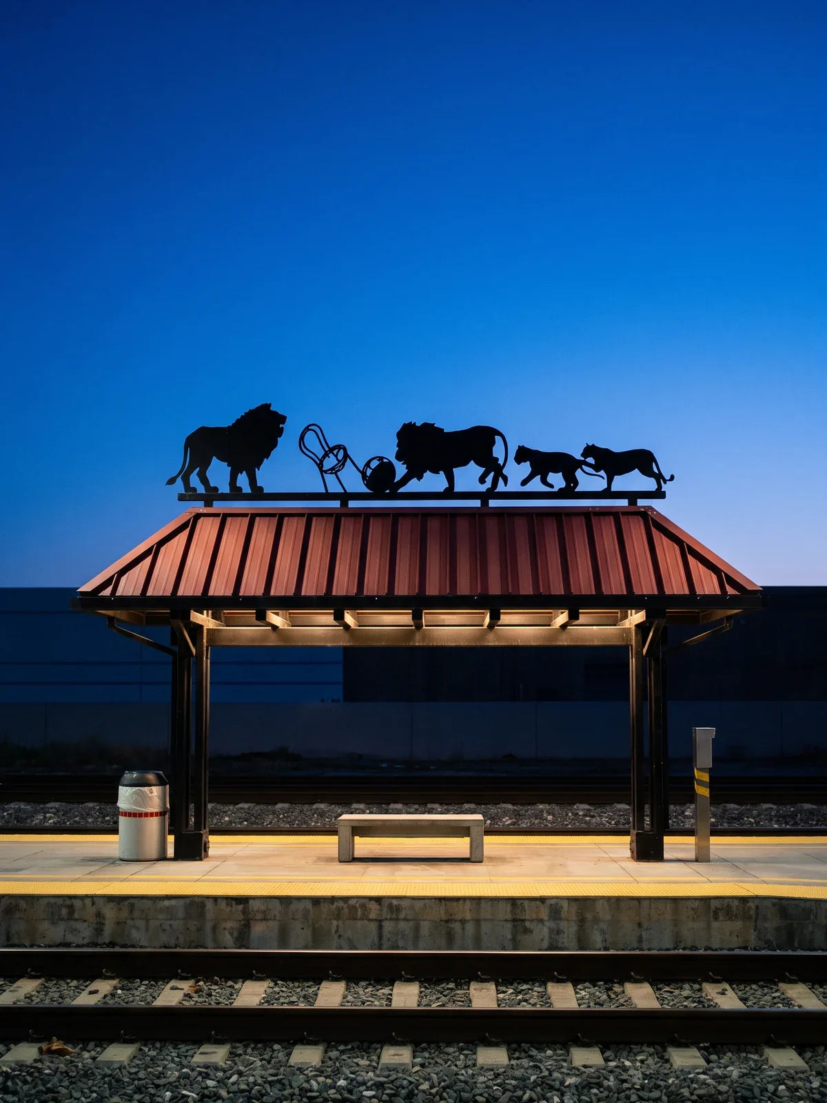 The lion canopy on the El Monte Metrolink platform at blue hour, black cut-metal lions doing tricks on a red-roofed shelter against a deep blue sky