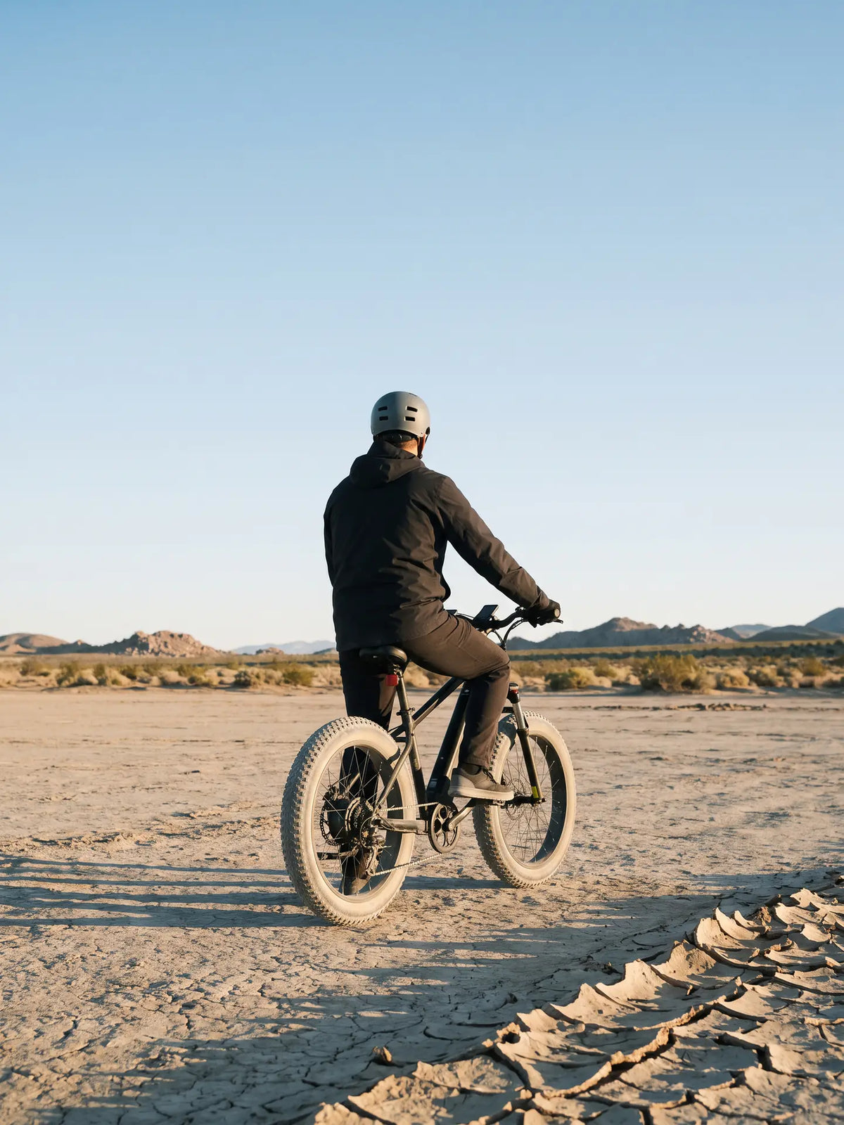 2000W e-bike buying guide cover: a rider on a fat-tire electric bike on a cracked dry lake bed, desert scrub and rocky hills behind.