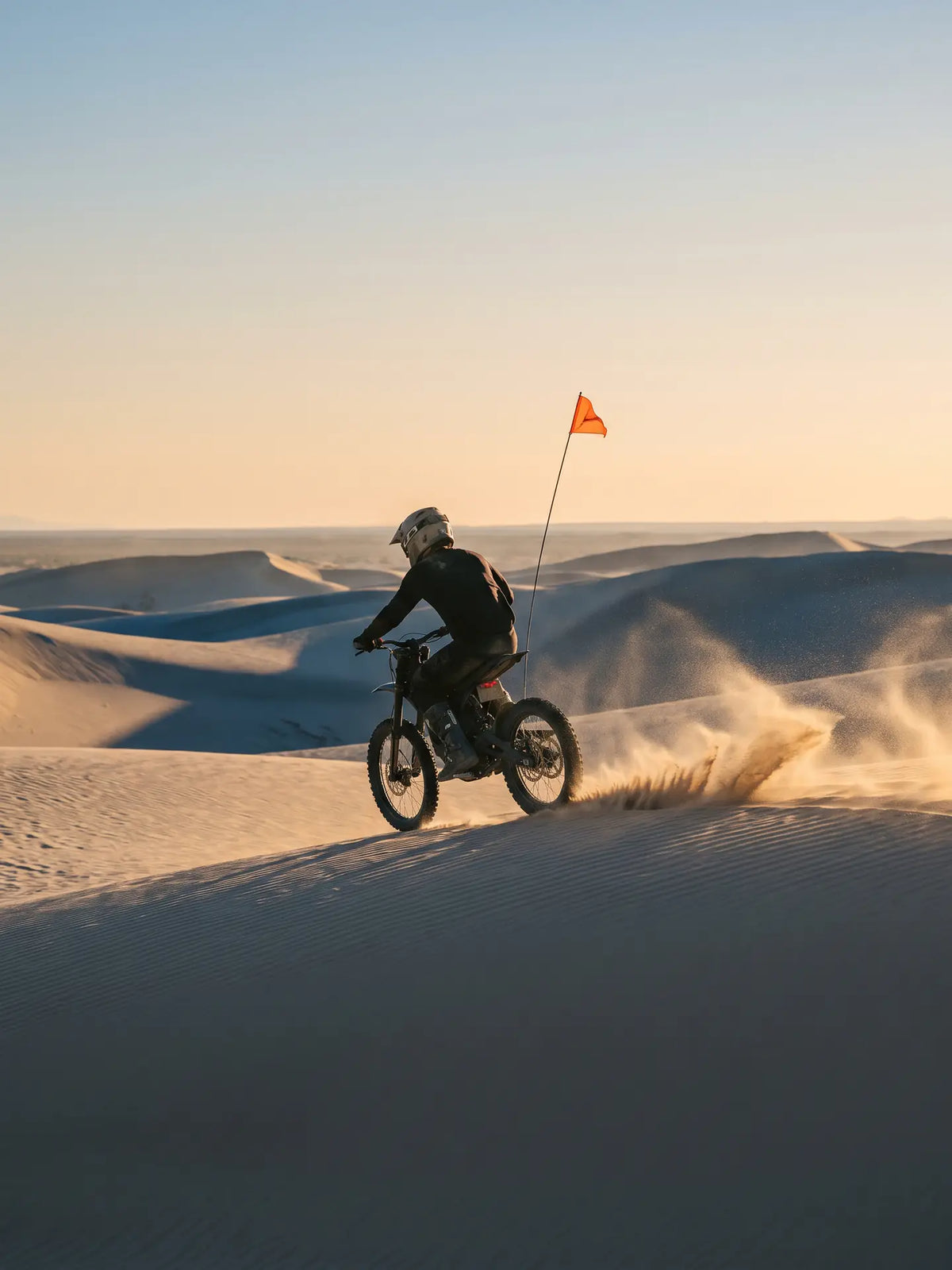 A 4,000W electric dirt bike cresting a sand dune at Imperial Sand Dunes Recreation Area in southeast California at golden hour, an orange safety flag on a whip mast behind it