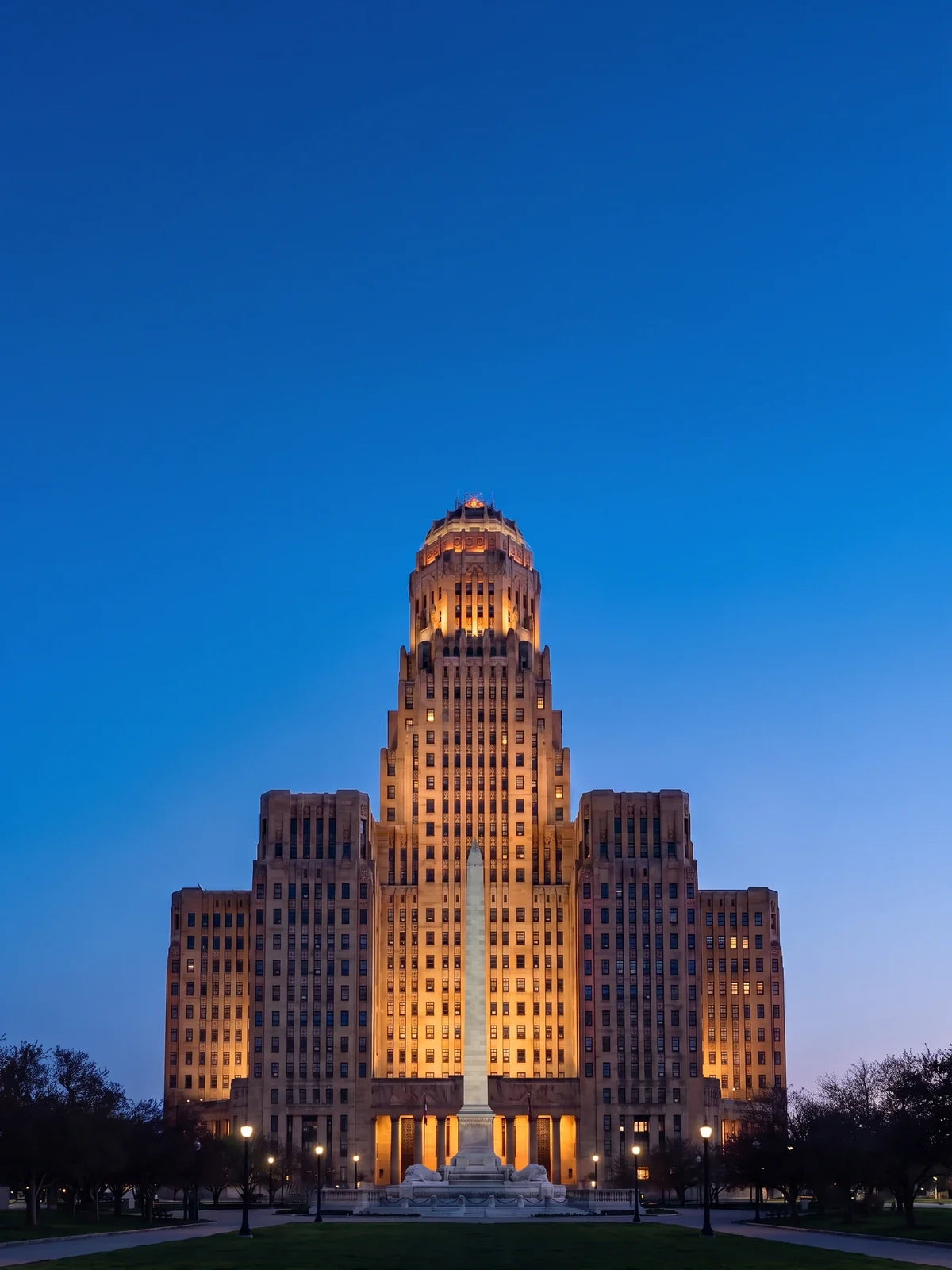 Buffalo City Hall at blue hour, an Art Deco tower of pale stone with a terra cotta crown, floodlit warm over Niagara Square, the McKinley Monument's white obelisk in front