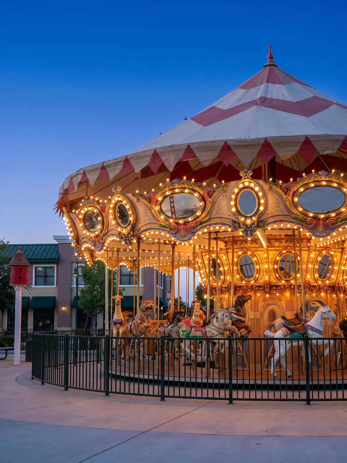 The carousel at Nut Tree Plaza at blue hour, its rows of small bulbs glowing warm along a red and cream canopy above carved animals behind a green fence