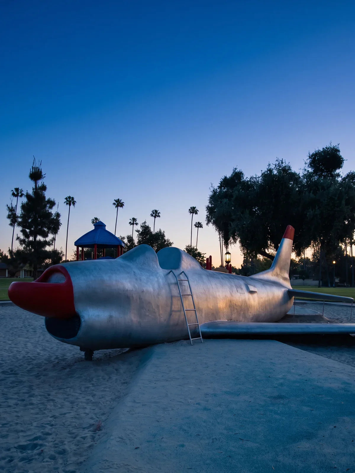 The retired Sabre jet on the playground at Palmview Park at blue hour, a silver swept-wing fighter with a red nose resting on the sand under a deep blue sky