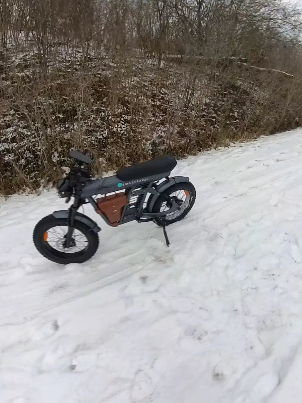 A gray Smartravel Rocket ST201F fat-tire e-bike with a brown frame bag, parked on its kickstand in fresh snow beside snow-dusted brush, one of this winter guide's 14 picks, on our own January ride