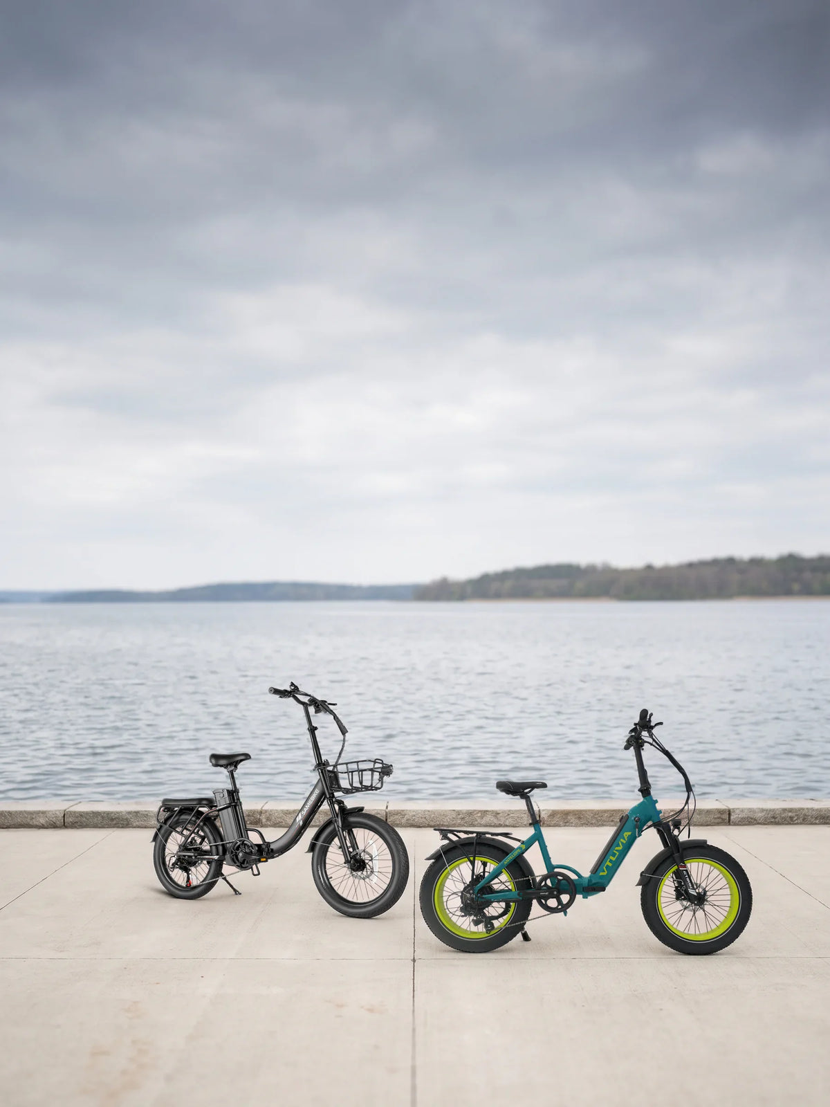 Lectric XP 3.0 review: two folding e-bikes we sell, the Samebike CY20 Pro in Black and the VTUVIA SX20 in Teal Blue, parked on a lakeside promenade under an overcast sky