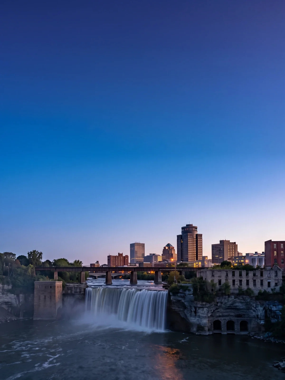 High Falls of the Genesee River at blue hour, the waterfall pale silver under the dusk sky, a low railroad bridge across its brink, a ruined stone mill on the gorge's rim and downtown Rochester's towers behind