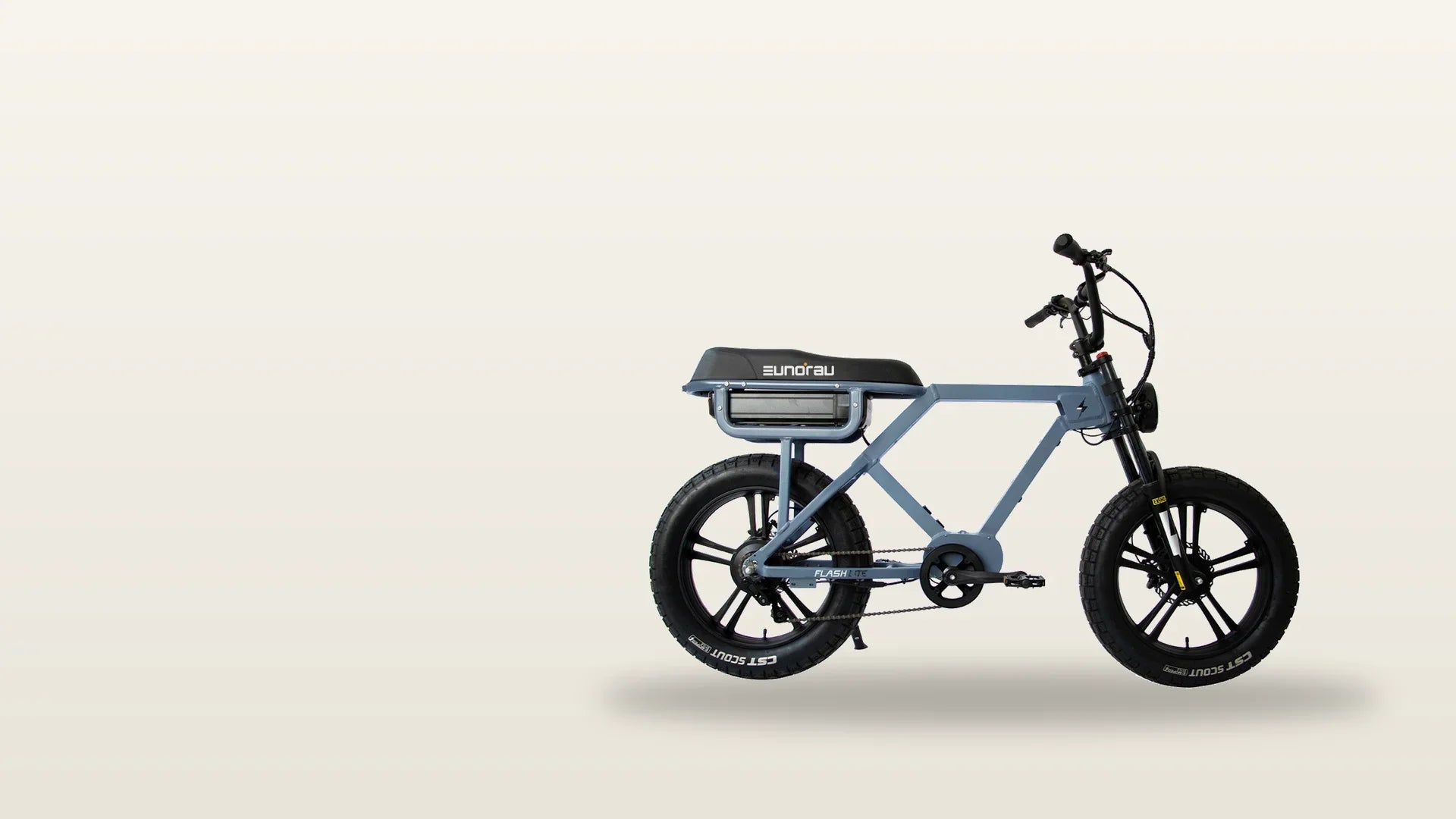 Rider in a denim jacket and half helmet leaning a moped-style electric bike through a wide city intersection at golden hour, bench seat and fat tires in profile