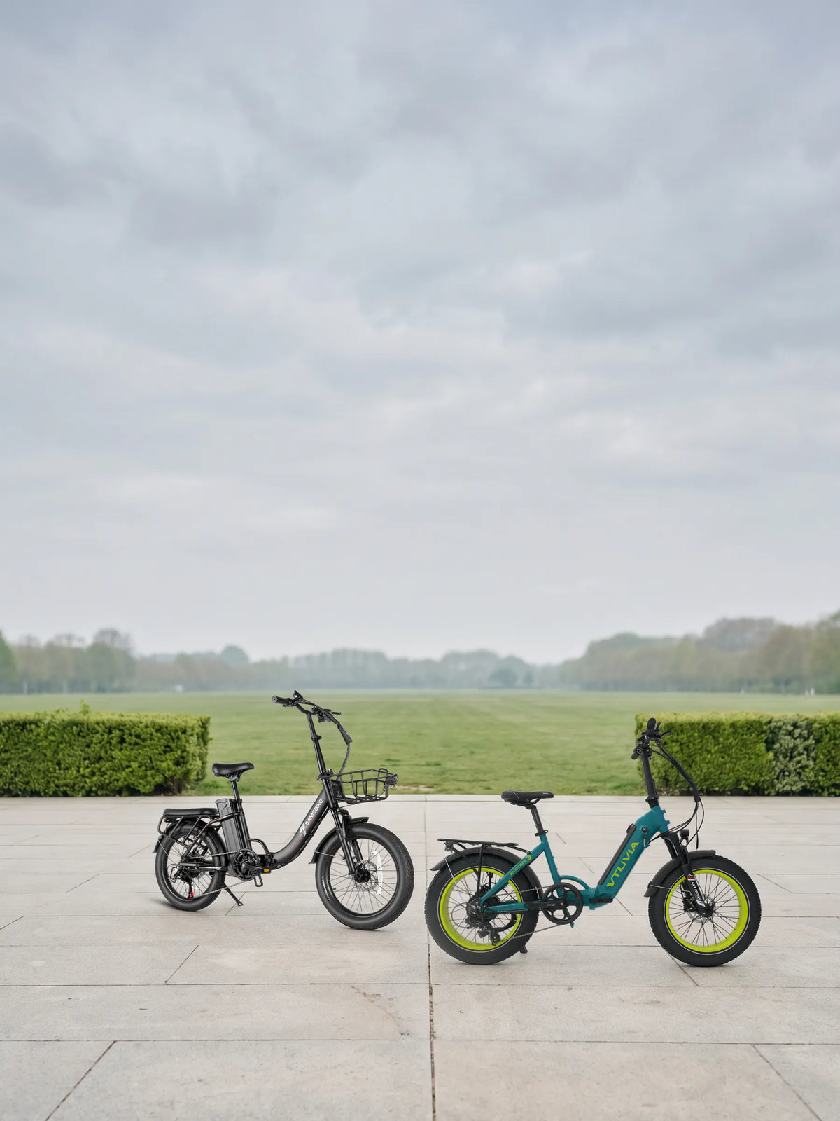 Lectric XP4 review: two folding e-bikes we sell, the VTUVIA SX20 in Teal Blue and the Samebike CY20 Pro in Black, parked on a paved park plaza under an overcast sky
