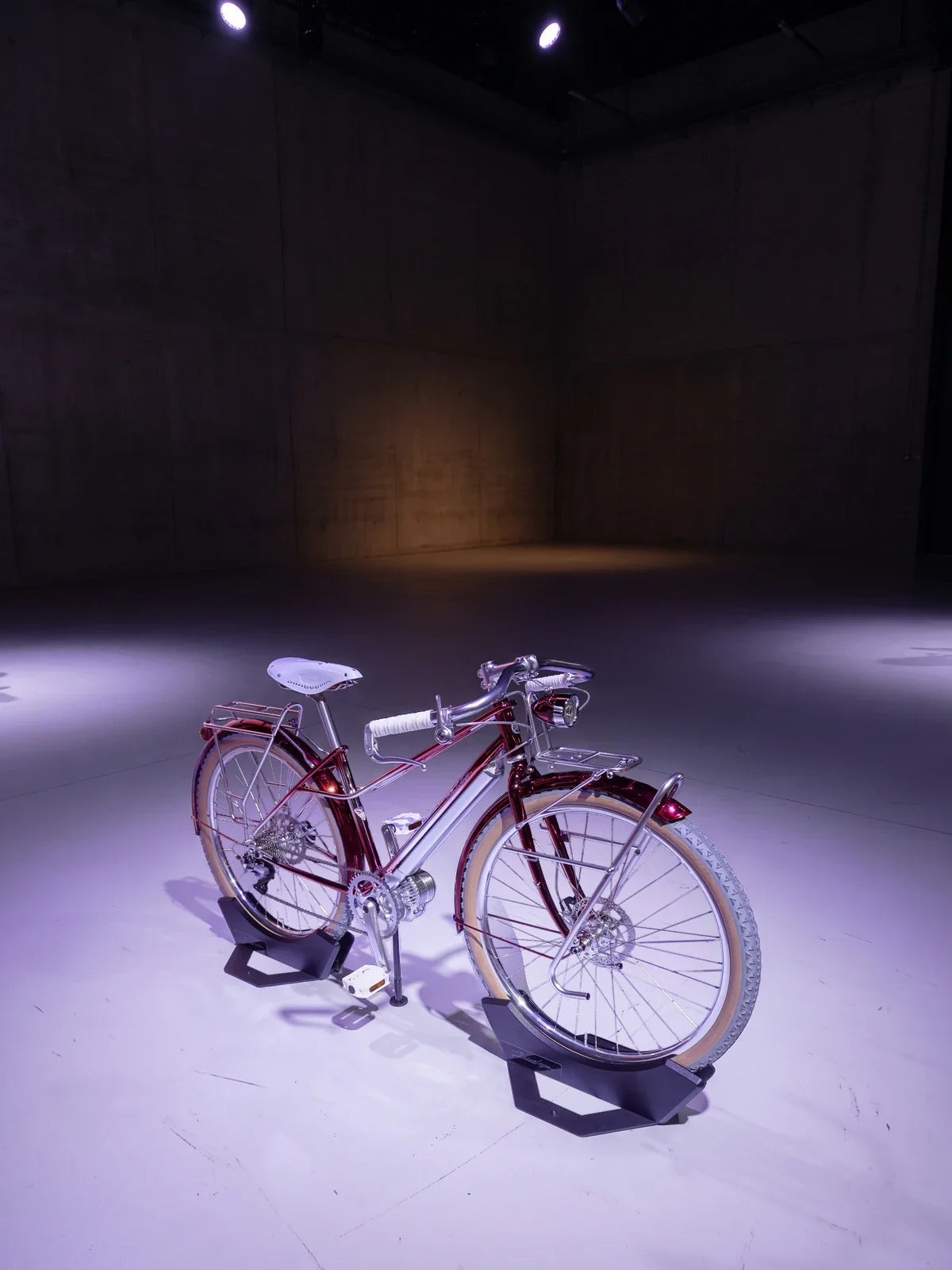 Yamaha's Y-00B: Bricolage concept e-bike, its candy-red frame and silver drive unit at the cranks lit violet-white, standing in two black display stands on the pale floor of a dark concrete hall