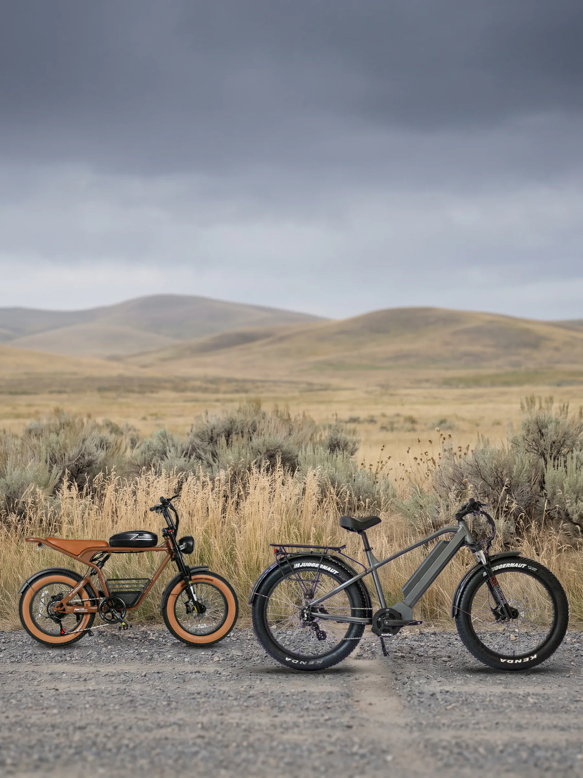 hunting e-bikes under $2,000: a Samebike M20 and a Eunorau FAT-HD 1.0 Pro parked on a gravel ranch road across open prairie under an overcast sky
