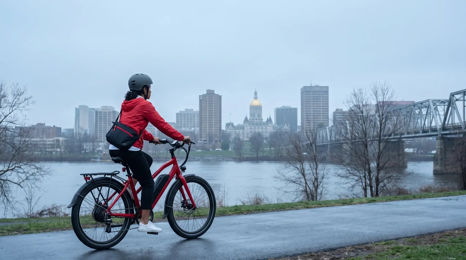 Cyclist in a helmet riding a red electric bike with a downtube battery on a paved riverfront path beside the Connecticut River, with the gold dome of the Connecticut State Capitol and the Hartford skyline behind